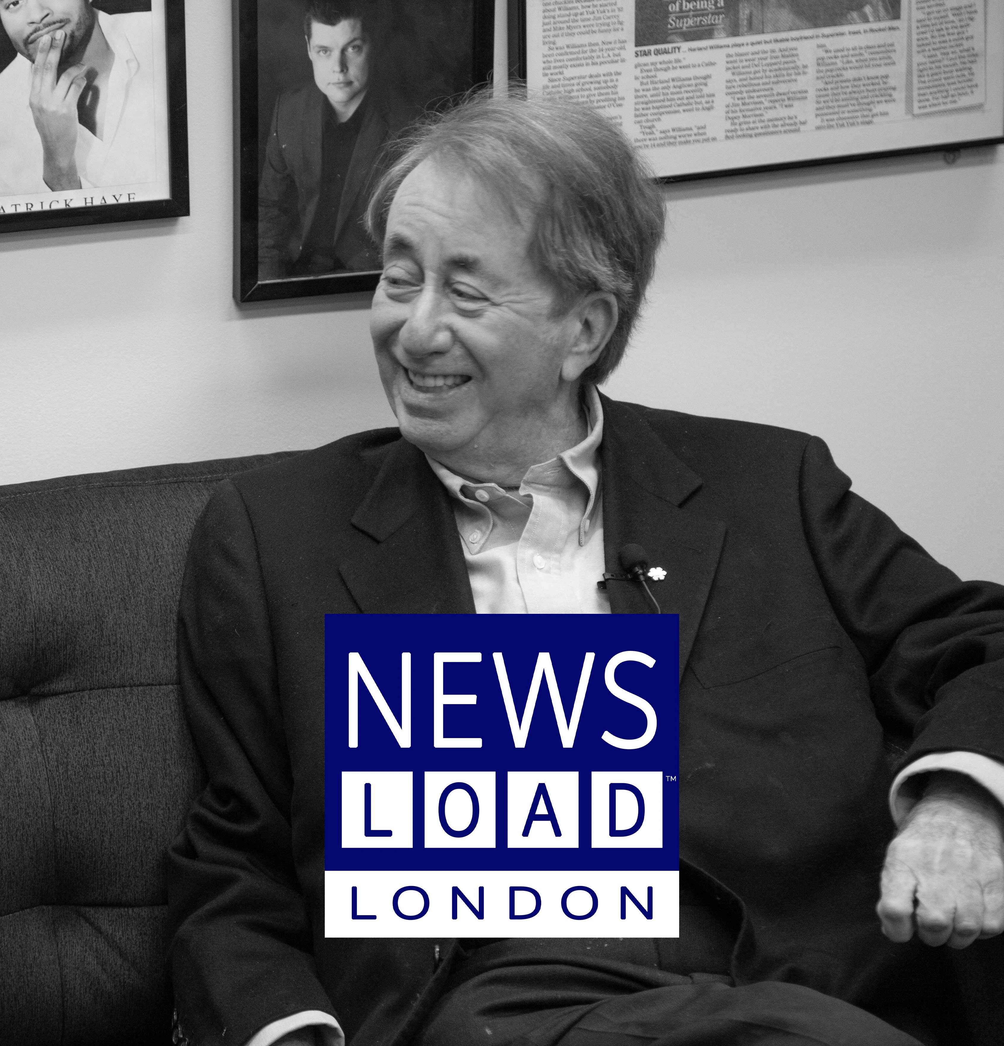 A man with light hair and a dark suit is seated on a dark couch, smiling and looking to the left, with framed pictures and text on the wall behind him. A blue and white logo with the words "NEWS LOAD LONDON" is superimposed over the lower center of the image.