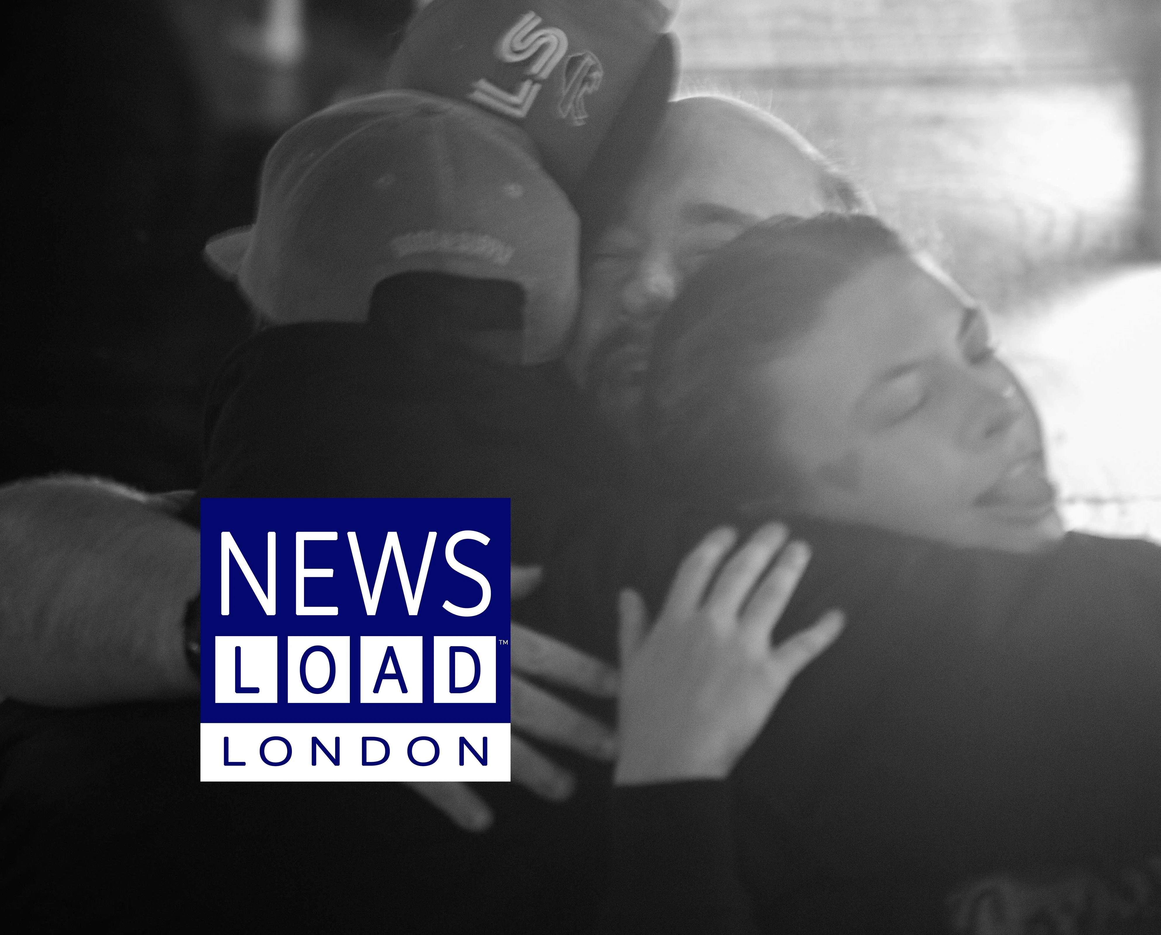 Three people are embracing in a dimly lit indoor setting, with one person wearing a baseball cap with "LS" visible on it, and another person's face partially visible with closed eyes.