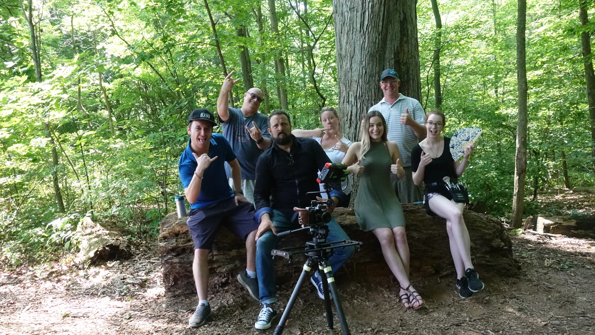 Seven people are posing for a picture in a forest, with a man in the center holding a camera on a tripod, and notable objects include a blue and white patterned fan and a water bottle.