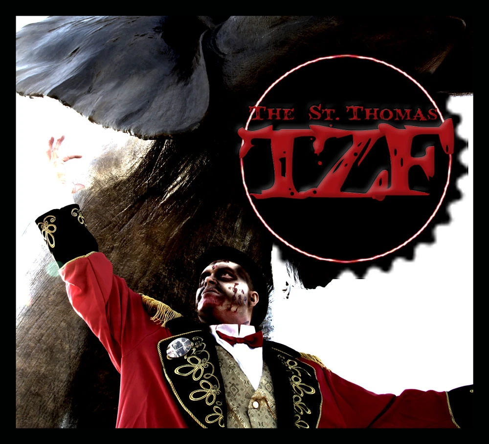 A person in a red and gold ringmaster's coat with white and red face paint is looking up with their right arm raised, standing next to a large, dark, textured object that resembles an elephant's ear and trunk, with a red circular logo containing the text "THE St. THOMAS TZF" in the upper right.