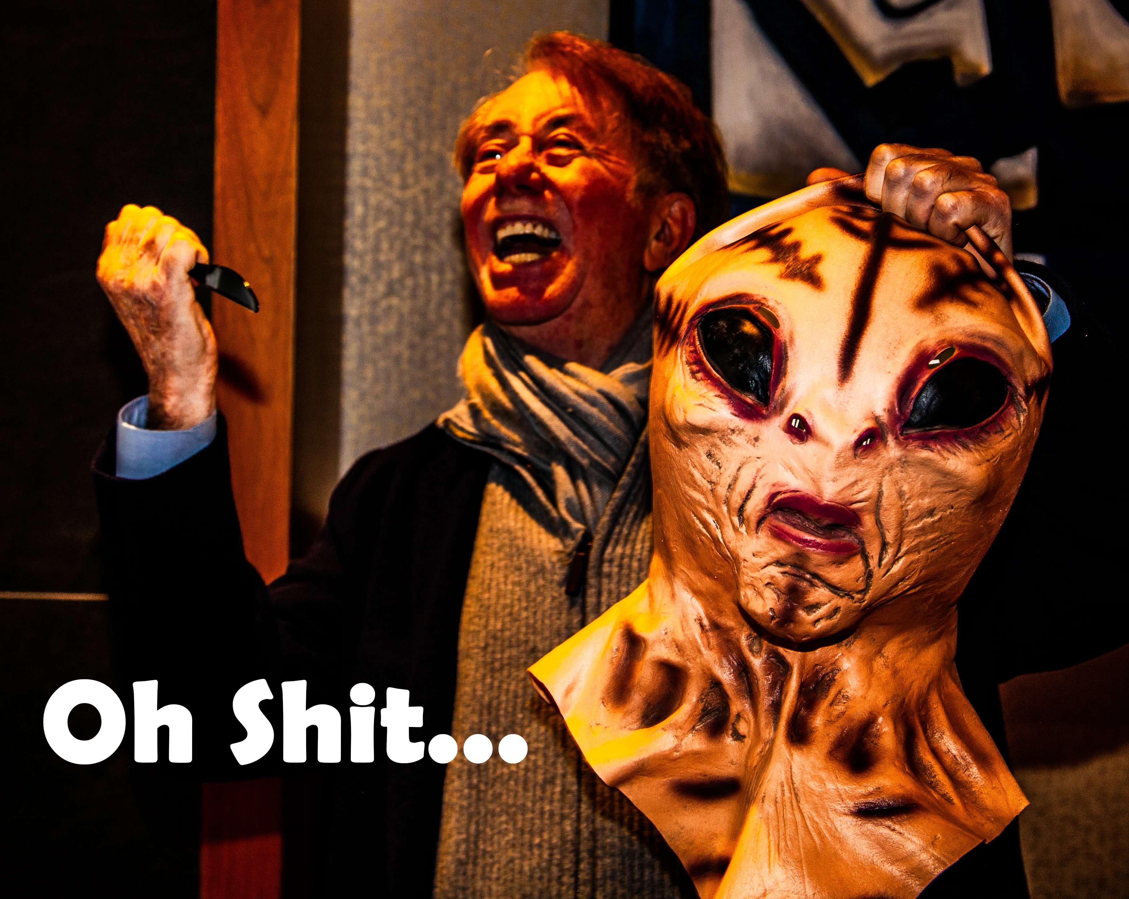 A man with short brown hair and a grey scarf is laughing with his mouth open, holding a black object in his raised left hand and an alien mask in his right hand, against a dark background with a brown wall on the right. The text "Oh Shit..." is visible in the bottom left corner.