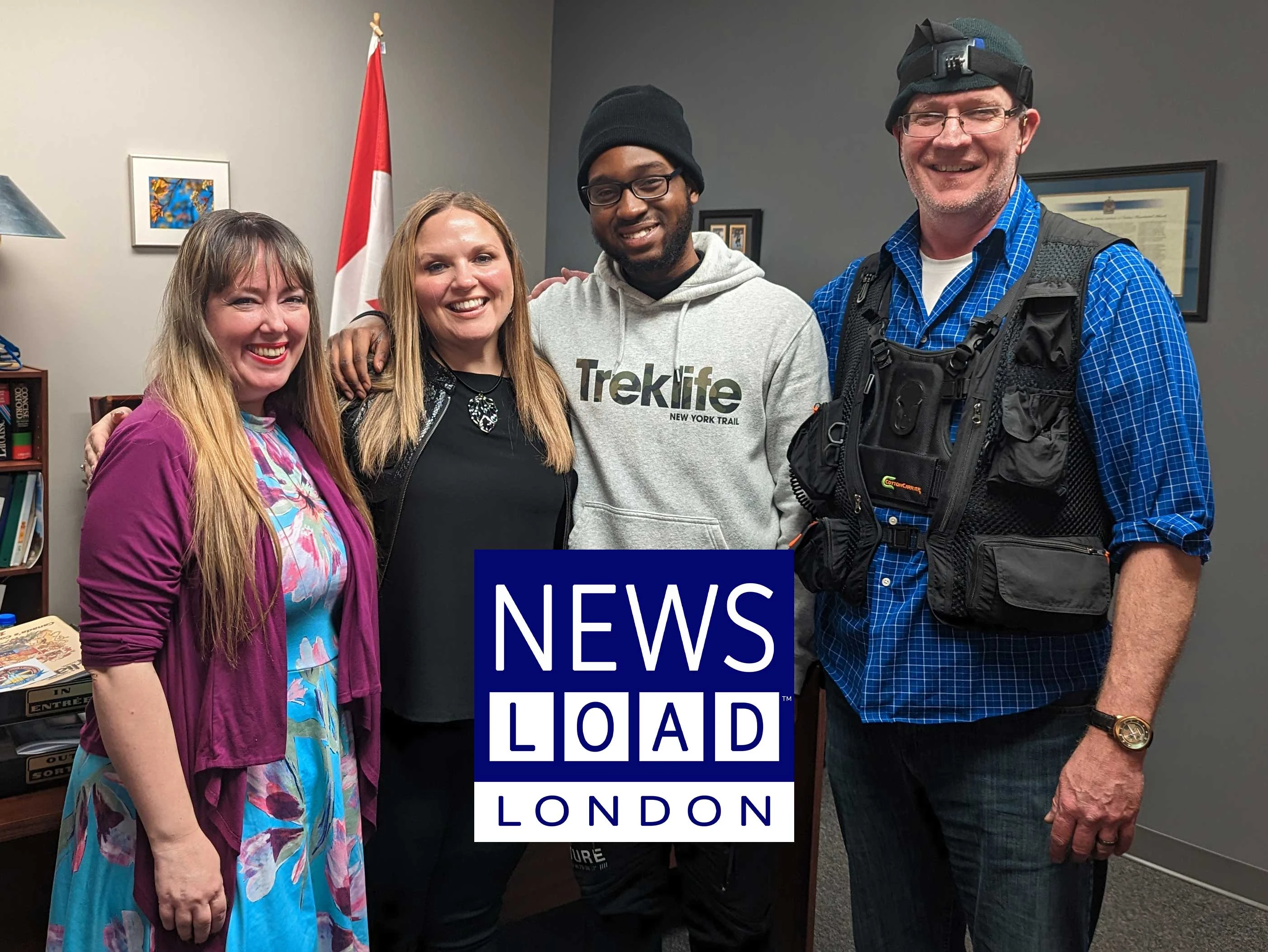 Four people are standing side-by-side, smiling at the camera, in an office with a Canadian flag, a bookshelf, and framed pictures on the wall. A blue and white graphic overlay with the words "NEWS LOAD LONDON" covers the lower center of the image.