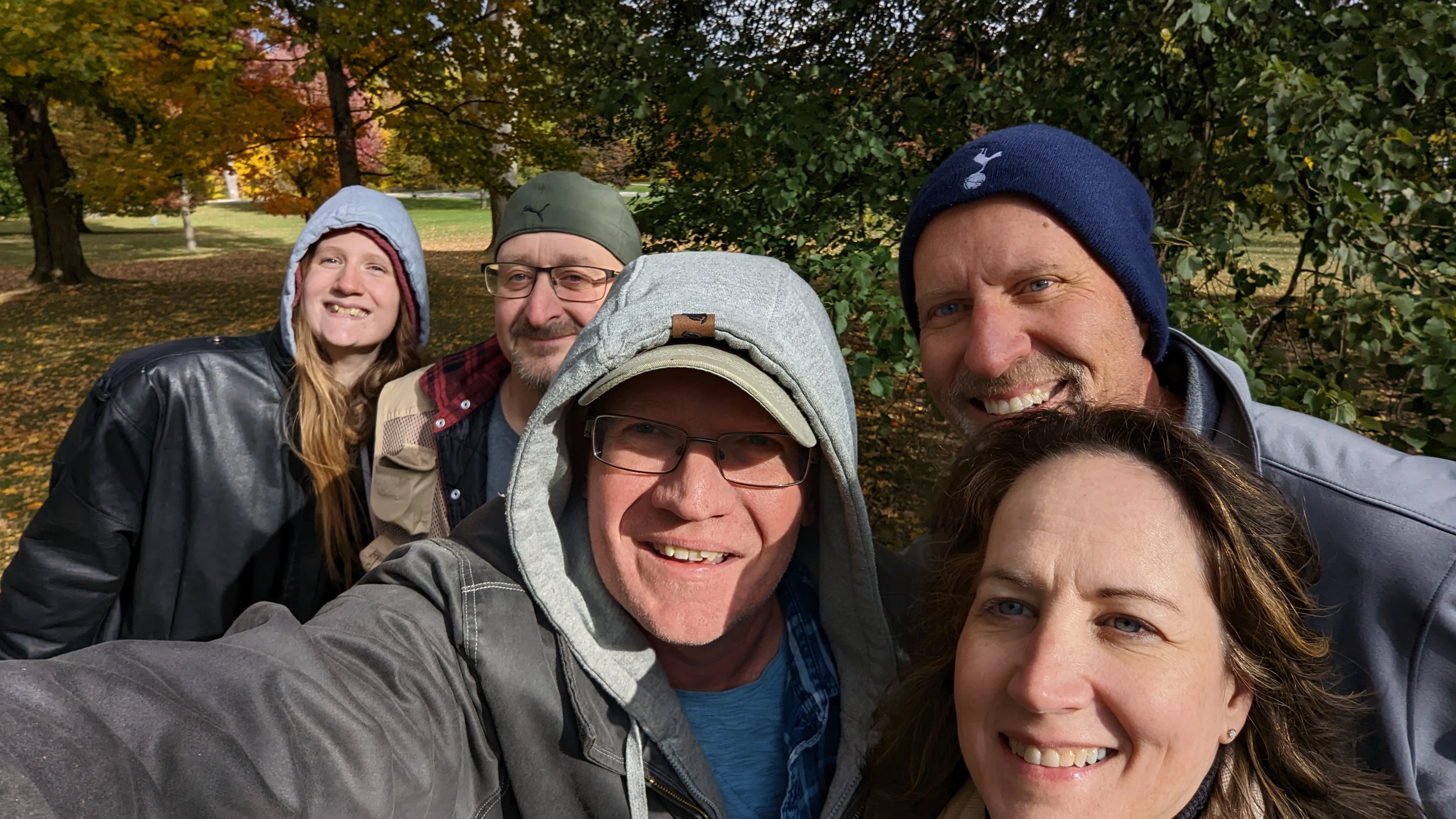 Five people are smiling for a selfie outdoors in a park with autumn trees, with one man wearing a Puma hat and another wearing a Tottenham Hotspur beanie.