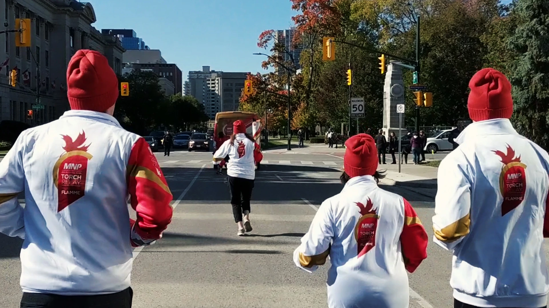 Three people wearing white jackets with red sleeves and red beanies are running on a city street, with one person in the middle running ahead of the other two, who are in the foreground with their backs to the viewer, on a sunny day with buildings and trees in the background, and several cars and traffic lights visible.