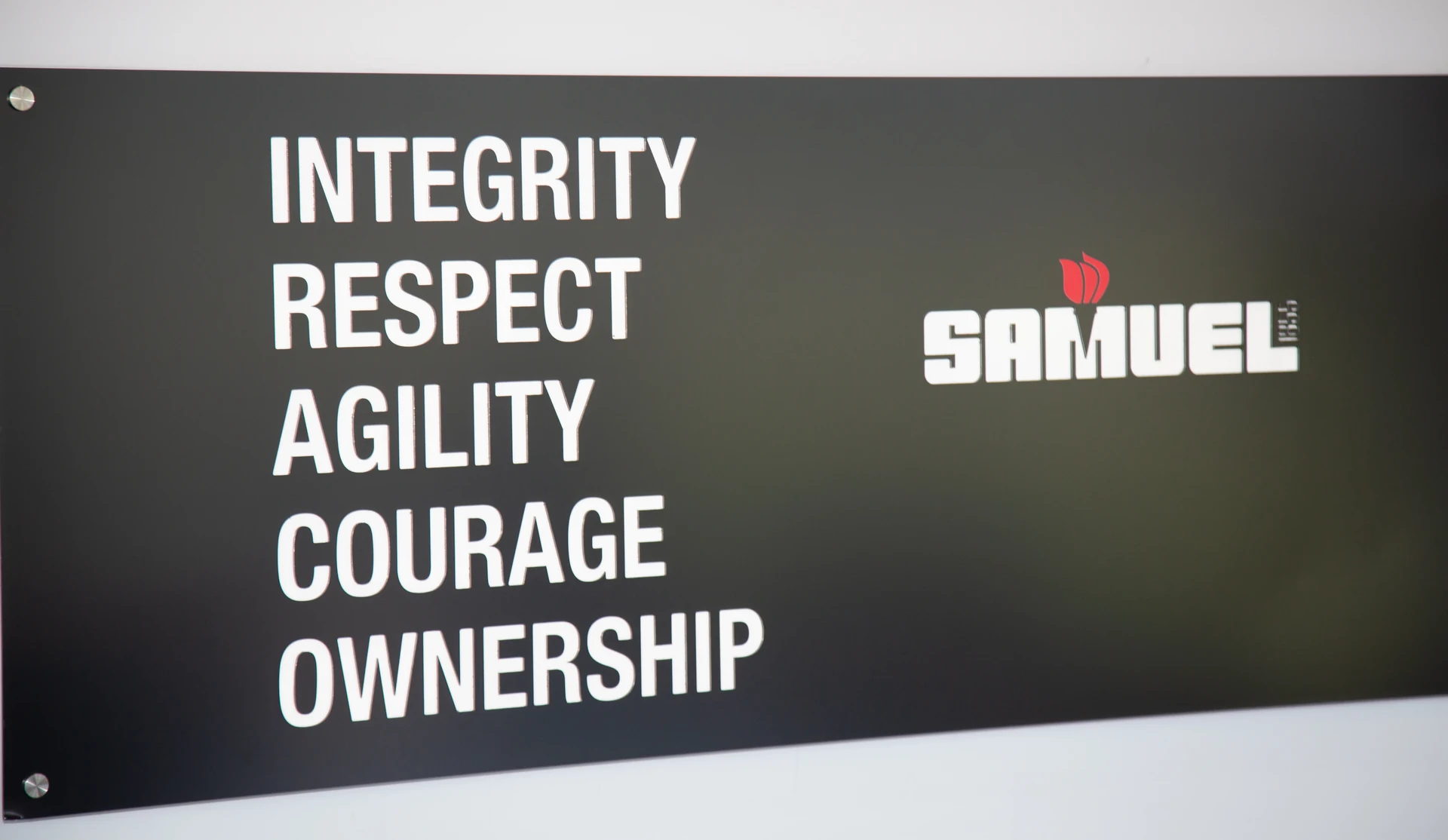 A black sign with white text is mounted on a white wall, displaying the words "INTEGRITY," "RESPECT," "AGILITY," "COURAGE," and "OWNERSHIP" on the left, and the "SAMUEL" logo with a red flame-like icon above it on the right.