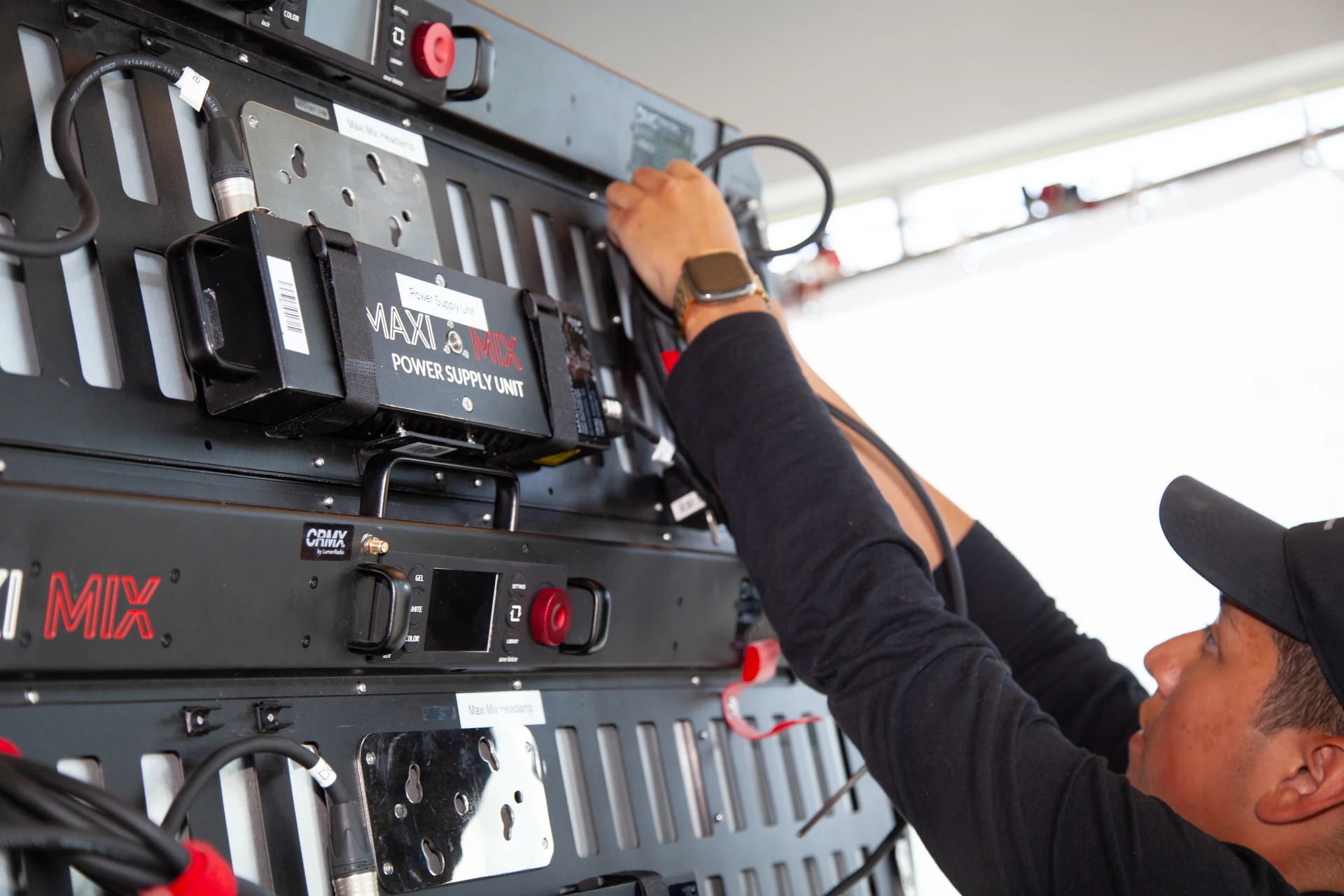 A person wearing a black long-sleeved shirt and a black baseball cap is connecting cables to a black equipment rack with various electronic components, including a "MAXI MIX POWER SUPPLY UNIT" and a "CRMX by LumenRadio" device.