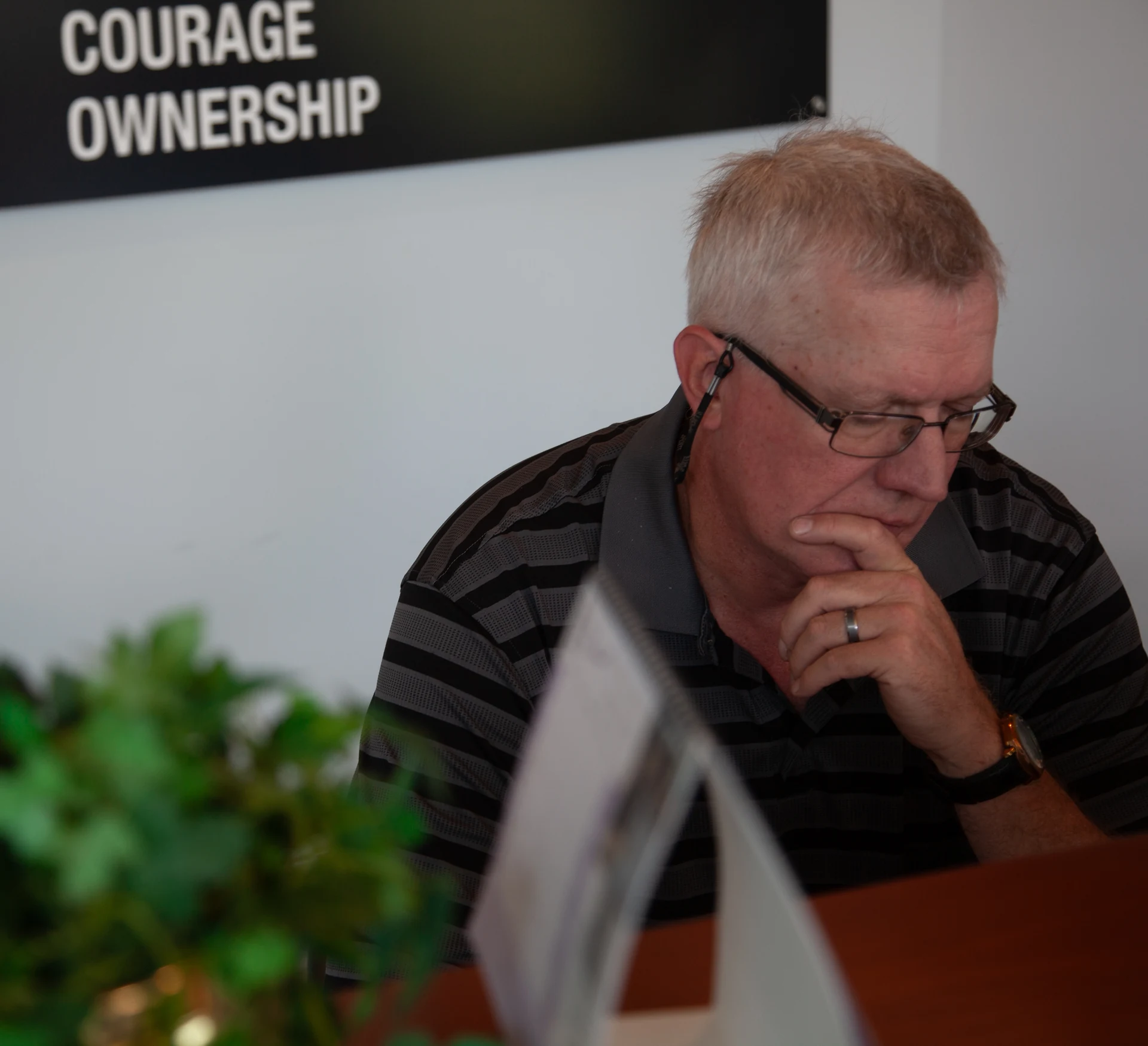 A man with short, light-colored hair and glasses is looking down, with his right hand touching his chin, wearing a striped polo shirt, a ring on his left ring finger, and a watch on his left wrist, in an indoor setting with a white wall and a black sign that reads "COURAGE OWNERSHIP" in white text, with a blurred green plant and a white folded paper object in the foreground.