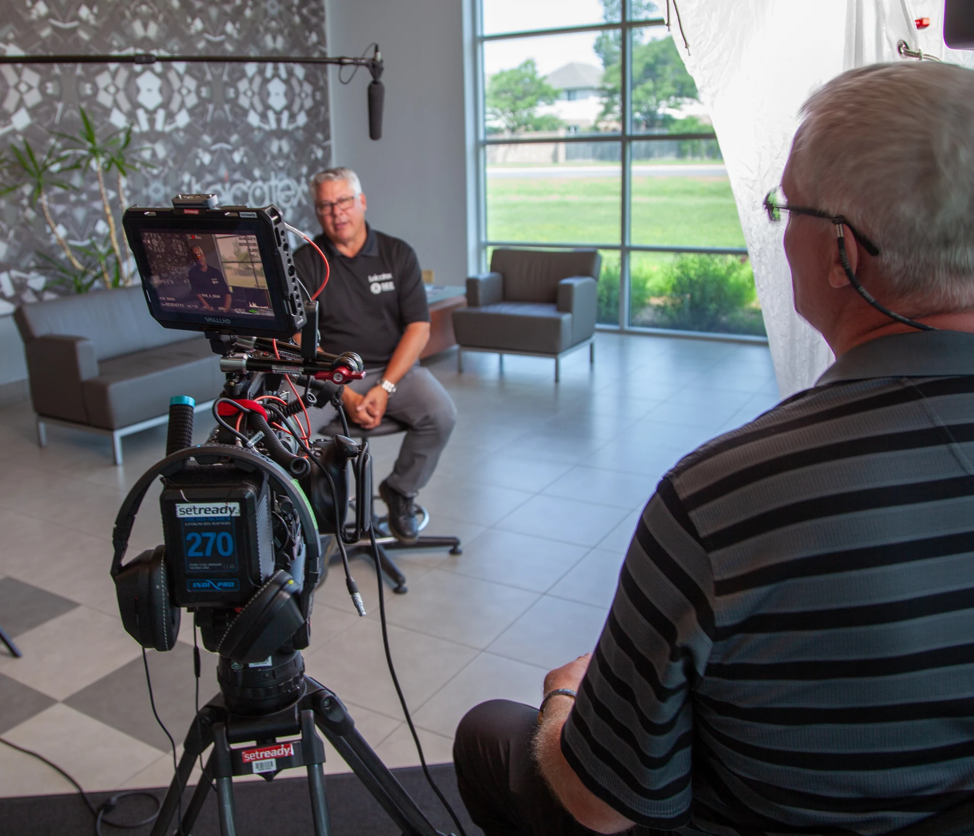 A man in a black polo shirt is being filmed by a camera on a tripod, with another man in a striped shirt sitting in the foreground, in a modern office setting with large windows and a patterned wall.