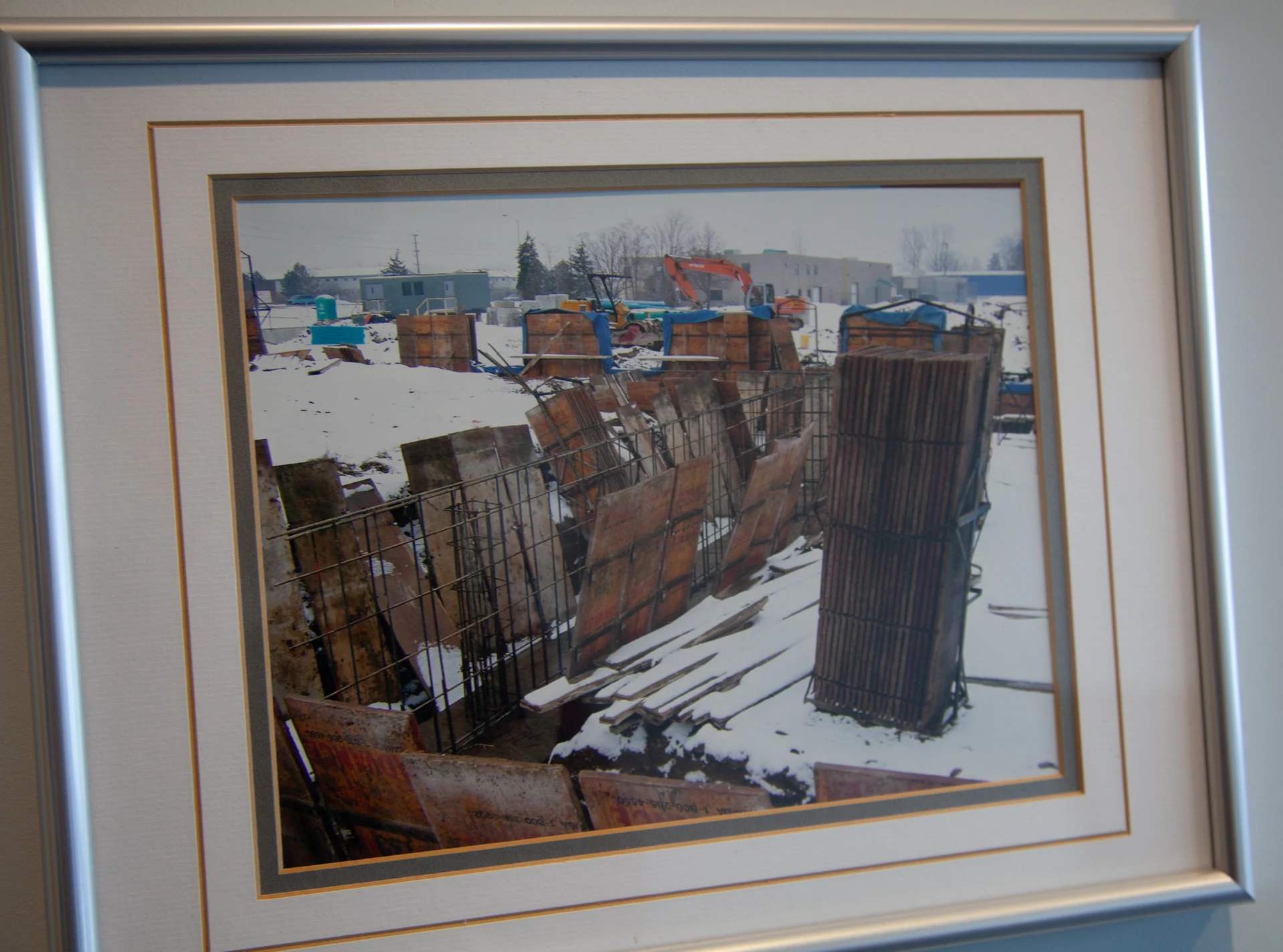 A framed photograph shows a construction site covered in snow, with various wooden forms, metal rebar, and construction equipment visible, including an orange excavator in the background.