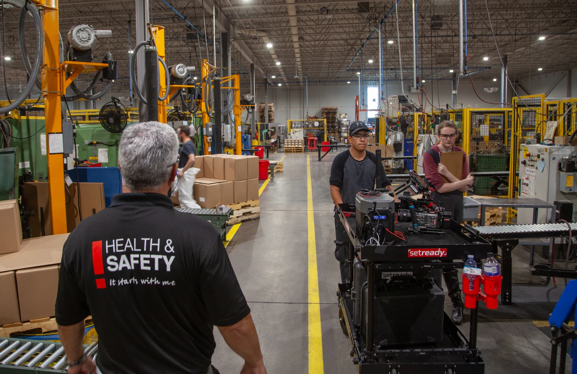 A man with gray hair wearing a black shirt with "HEALTH & SAFETY It starts with me" printed on the back is walking away from the viewer in a large industrial warehouse with yellow safety lines on the floor, while another man in a black cap and glasses pushes a black cart with camera equipment and a woman in glasses holds a clipboard.