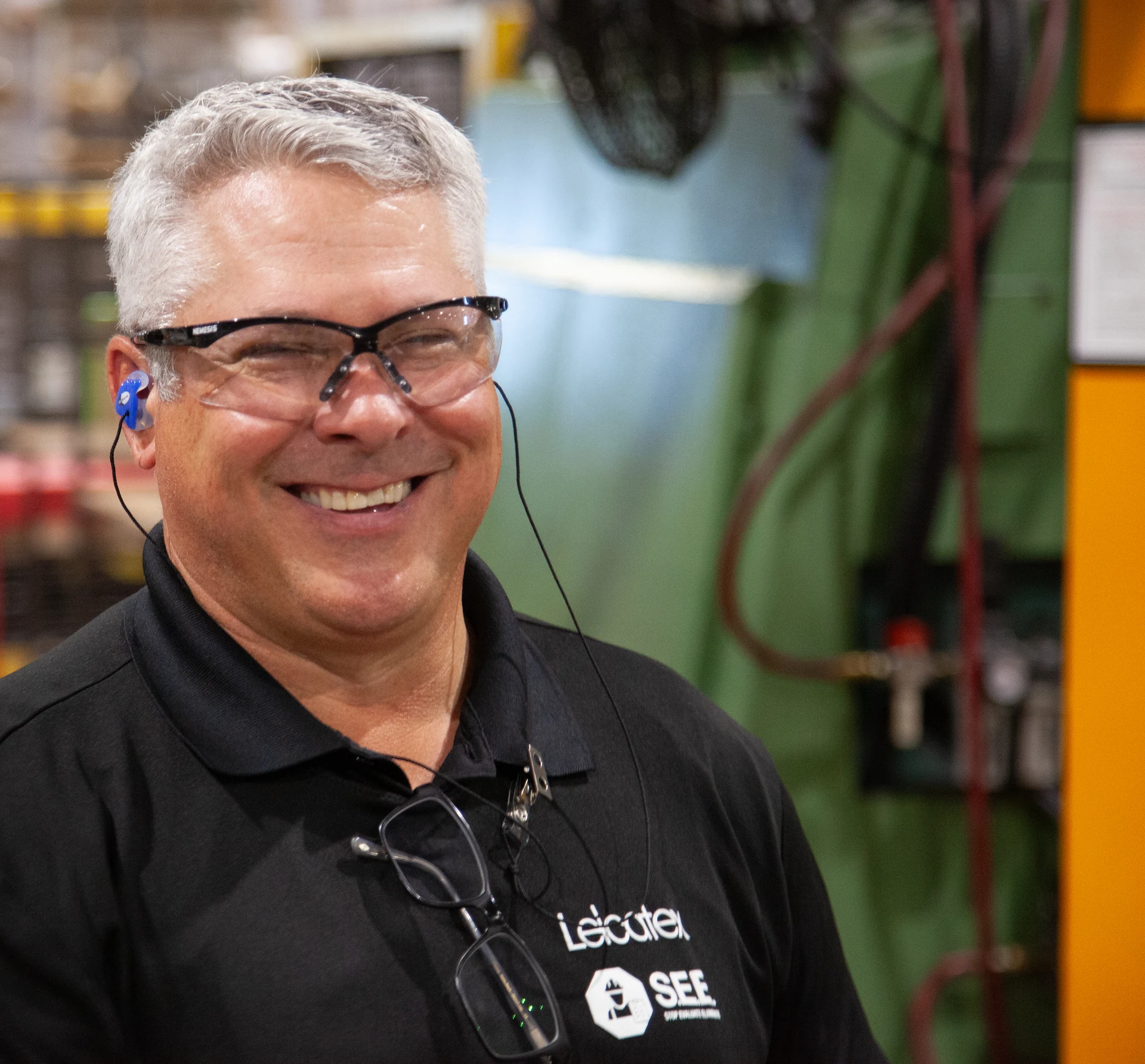 A man with gray hair and safety glasses is smiling, wearing a black polo shirt with a lanyard holding eyeglasses, and blue earplugs, in what appears to be a factory or workshop setting with machinery and pipes in the background.