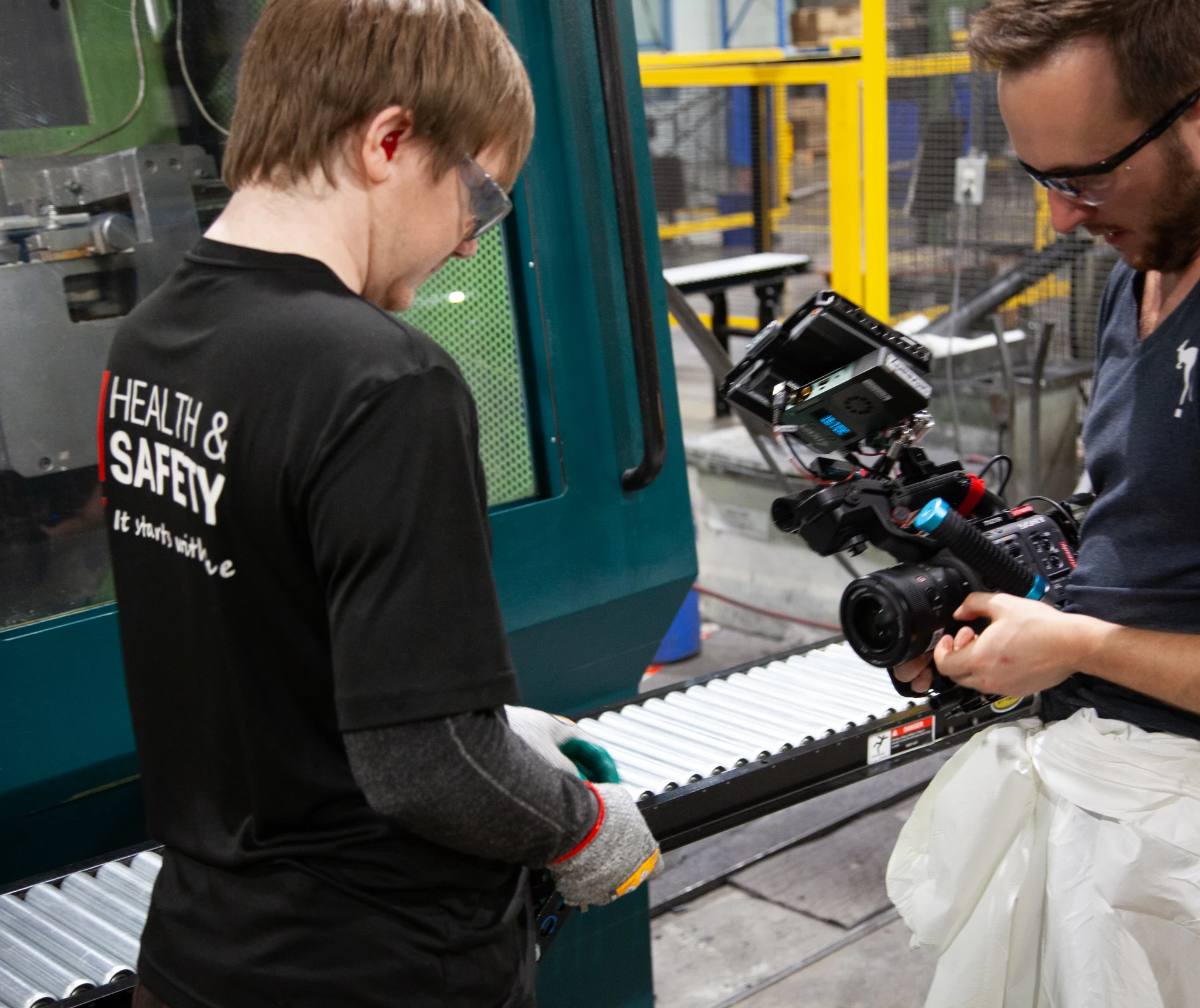A man wearing a black shirt with "HEALTH & SAFETY It starts with us" printed on the back, safety glasses, and earplugs is standing next to a teal machine with a roller conveyor, while another man in a blue shirt and glasses holds a camera with a monitor attached.