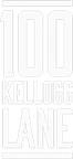The main subject is a white sign with the text "100 KELLOGG LANE" in a bold, outlined font with two horizontal lines above and below the text.