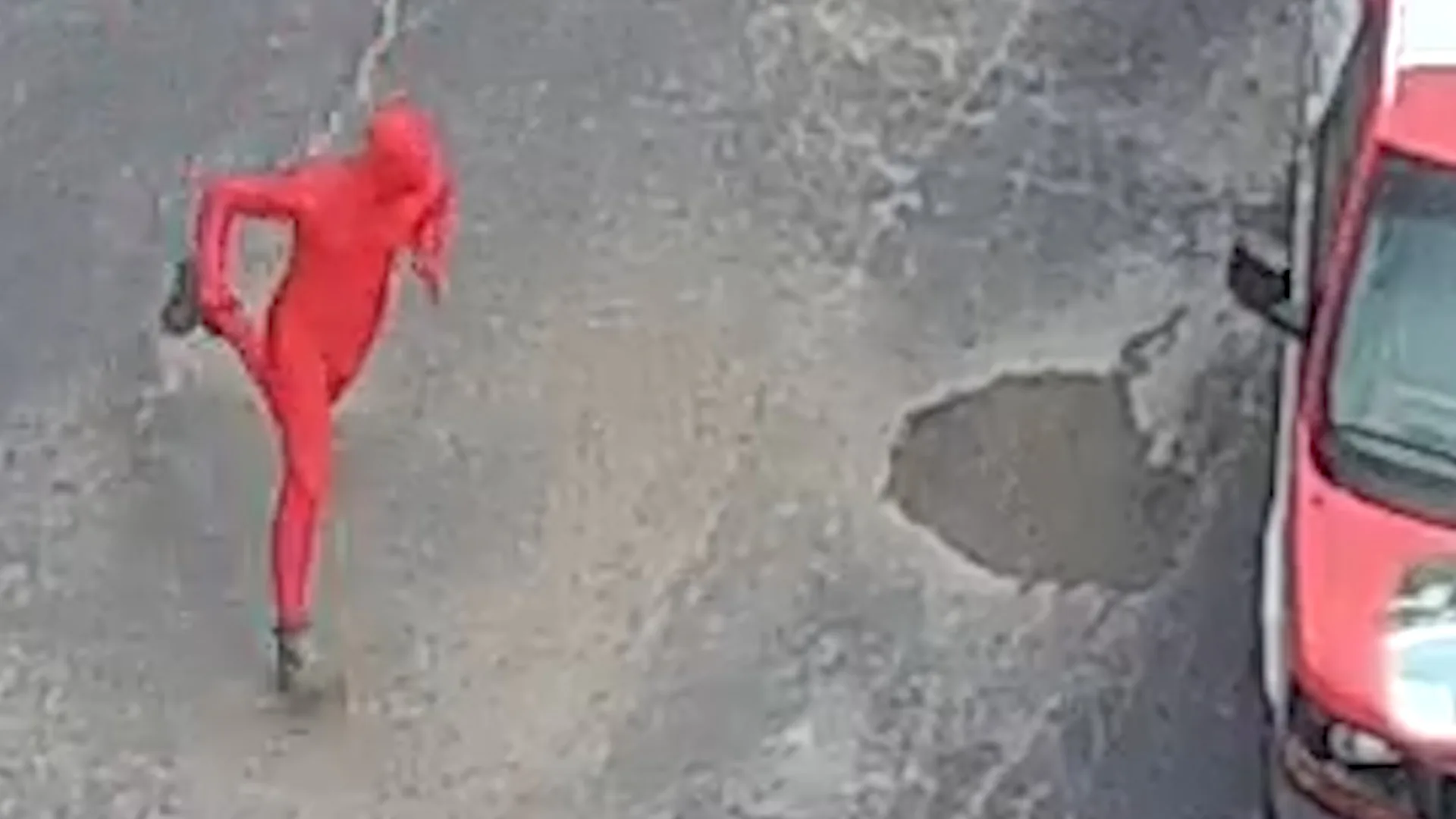 The mascot, on location. An edited still — the red-suited figure outdoors on a road, a red vehicle at the edge of frame. A frame from the series, not a photo of the making of it.