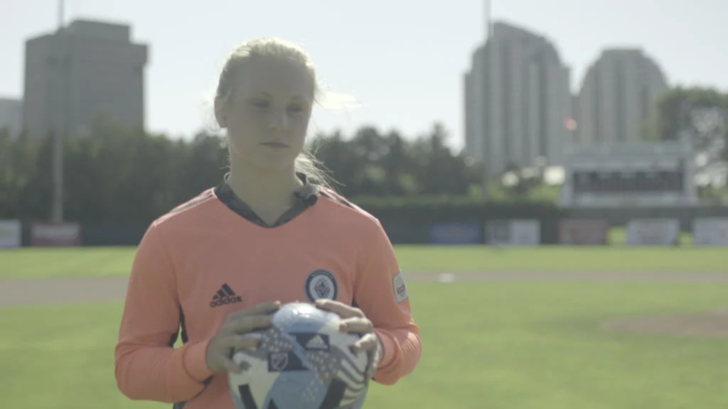 A female soccer player in a peach long-sleeved jersey with black trim holds a white, blue, and black soccer ball with both hands in front of her on a green sports field, with blurred buildings and trees in the background under a bright sky.
