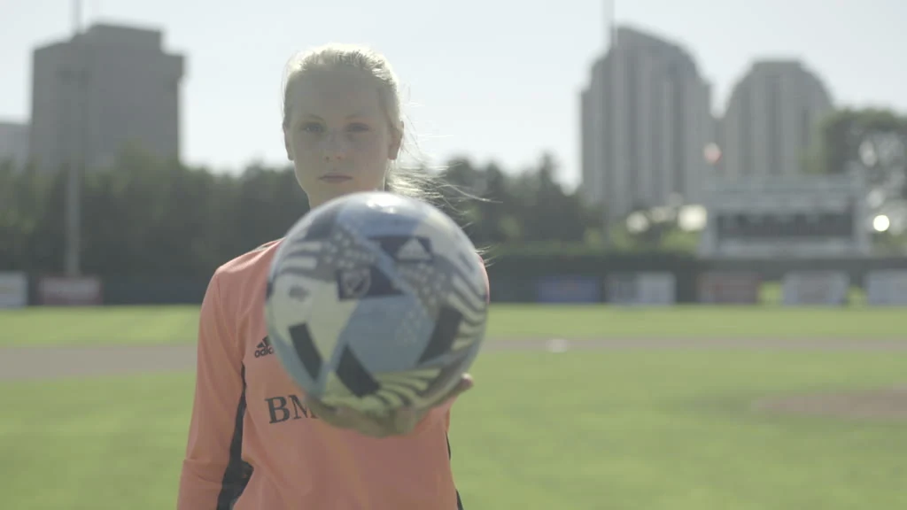 A young woman in an orange long-sleeved shirt holds a soccer ball in front of her on a sunny day at a sports field with buildings in the background.
