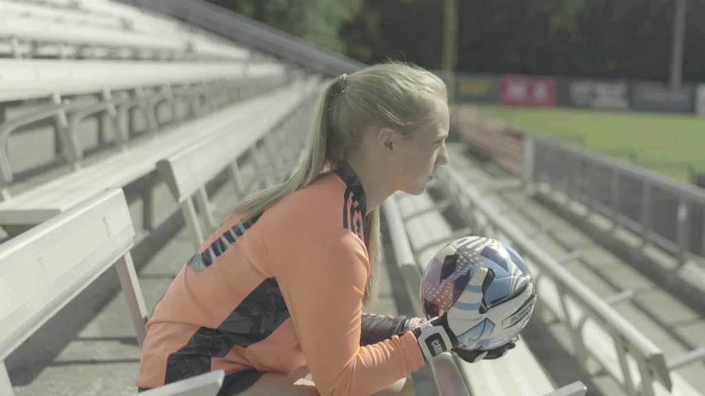 A blonde woman in an orange and black long-sleeved shirt and black shorts sits on a white bleacher, holding a blue, white, and black soccer ball with both hands, wearing white and black gloves, and looking to the right, with more bleachers and a green field visible in the background.