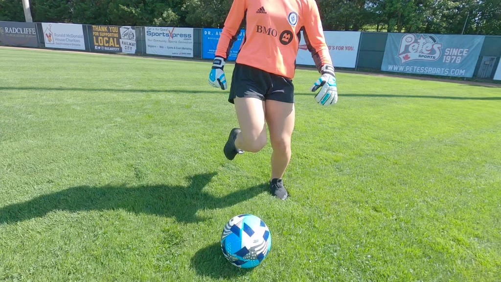 A person wearing an orange long-sleeved shirt, black shorts, and white and blue gloves is standing on a green grass field with a blue, white, and black soccer ball in front of them, with advertising banners visible in the background including "RADCLIFFE", "Ronald McDonald House Charities Southwestern Ontario", "THANK YOU FOR SUPPORTING LOCAL", "RAILWAY CITY", "SportsXpress", "BMO", and "Pete's SPORTS SINCE 1978 WWW.PETESSPORTS.COM".