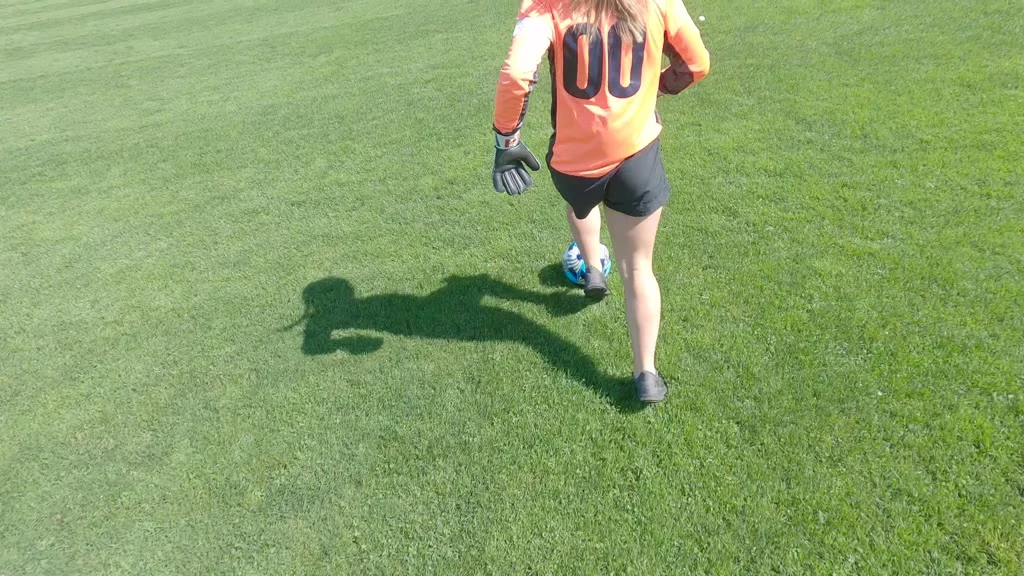 A person wearing an orange jersey with "00" on the back, black shorts, and black gloves is kicking a blue and white soccer ball on a green grass field, with their shadow visible on the grass.